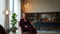 Professional businesswoman in burgundy suit sitting confidently in modern office lobby with contemporary lighting and plants for corporate branding.