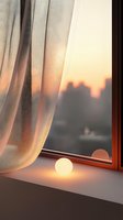Glowing sphere lamp on windowsill at sunset with sheer curtain and city silhouette view creates cozy atmospheric mood for interior design and lifestyle concepts.