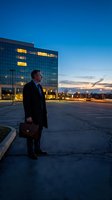 Professional businessman in dark suit holding briefcase stands outside modern office building during blue hour twilight for corporate success concepts.