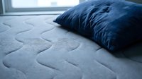 Close-up of a blue pillow resting on a quilted mattress surface with stitched patterns, showing bedroom comfort and sleep setup for home decor and wellness content.