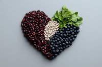 Heart-shaped composition of red kidney beans, white navy beans, and fresh blueberries with green lettuce leaves on gray background, promoting healthy eating.