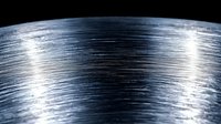Metallic brushed steel surface with linear texture and light reflection creating dynamic industrial pattern. Macro photography reveals detailed scratched finish.