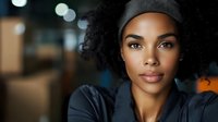 Young African American woman with curly hair wearing gray beanie and hoodie looking at camera with confident expression in urban indoor setting with bokeh lights.