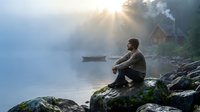 Young bearded man sitting on rocky shore contemplating misty lake at sunrise with wooden boat and cabin creating peaceful morning meditation scene.