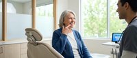 Senior Caucasian woman with gray hair smiling during dental consultation with male dentist in modern clinic office with natural lighting and professional equipment.