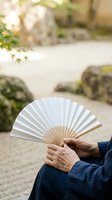 Senior woman holding white and natural wood folding fan in zen garden with raked sand and moss-covered stones, peaceful outdoor setting for relaxation and wellness concepts.