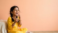 Young South Asian girl in bright yellow shirt sitting against warm peach background, laughing joyfully while holding microphone, expressing happiness and enthusiasm for singing or performance activities.