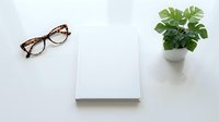 Blank white notebook with eyeglasses and green plant on clean desk workspace for business planning and creative writing projects.