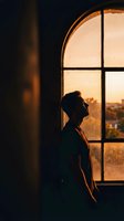 Young man silhouette gazing through arched window at golden sunset cityscape, contemplative mood in warm interior lighting for lifestyle concepts.