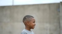 Young Black male with short hair and geometric line tattoo on head, wearing gray shirt, standing in profile against concrete wall background.
