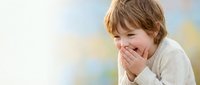 Young boy with tousled brown hair covering mouth while laughing joyfully against soft blurred background. Candid moment of childhood happiness and playful expression.