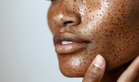 Close up view of dark skin with scattered freckles across cheeks and nose, glossy nude lips, and gentle touch of fingers against face showing natural beauty and skincare.