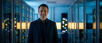 Male IT professional in dark business suit smiling confidently in modern data center with illuminated server racks, representing technology expertise, network infrastructure, and digital innovation.