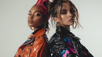 Two fashion models with dramatic makeup wearing colorful leather jackets and statement accessories against neutral background, showcasing high fashion editorial style.