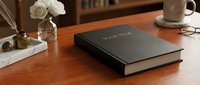 Black hardcover book with gold title text resting on a wooden desk beside a white rose, quill pen, and coffee mug in a cozy reading setup.