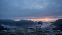 Wooden chair sitting alone on misty mountain landscape at dawn, surrounded by fog-covered peaks and soft golden sunrise light breaking through clouds.