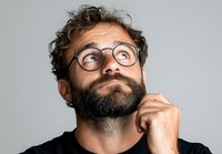 Thoughtful bearded Caucasian man with glasses looking up while thinking, wearing black t-shirt against light background. Contemplative facial expression shows curiosity.