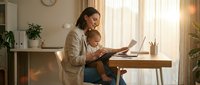 Young professional mother holding infant baby while working on laptop at home office desk during golden hour sunlight streaming through window.