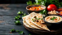 Middle Eastern appetizer spread with fresh pita bread, creamy hummus dip, tomatoes and chickpeas on rustic wooden board against dark background. Food photography.