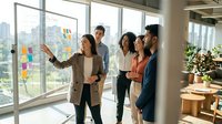 Woman in beige blazer leading diverse team brainstorming session with sticky notes on glass wall in modern office with city skyline view.