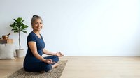 Mature woman with gray hair and light skin tone practicing meditation in lotus position on yoga mat indoors, wearing navy blue shirt, suitable for wellness and mindfulness content.