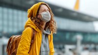 Woman with wavy hair wearing protective face mask, yellow coat and beanie at airport terminal. Traveler with backpack during pandemic.