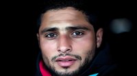 Young Middle Eastern man with facial hair and intense gaze looking directly at camera against dark background, dramatic portrait lighting emphasizes facial features.