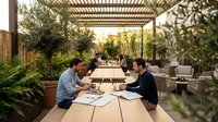 Two professional businessmen having outdoor meeting on wooden deck terrace with pergola discussing documents and paperwork in modern garden setting.