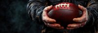 Dramatic close-up of hands holding brown leather football against dark background with atmospheric smoke effect, capturing intense sports moment in panoramic view.