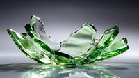 Shattered green glass fragments arranged in artistic formation against gray background, creating dynamic sculptural effect with reflective surfaces and sharp edges.