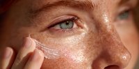Young Caucasian woman applying white moisturizing cream on freckled face skin during daily skincare routine for healthy glowing complexion care.