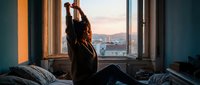 Woman with long reddish-brown hair, sitting on bed by window at sunrise, stretching arms up, wearing dark clothing, urban cityscape visible in background.