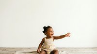 Young African American toddler girl in patterned outfit sitting on wooden floor with arm extended, expressing joy and playfulness against neutral background for childcare and parenting content.