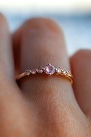 Delicate rose gold engagement ring with pink sapphire center stone and small diamond accents shown on finger against soft blurred background.