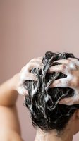 Woman washing dark curly hair with shampoo foam in shower against pink background for haircare and beauty routine.