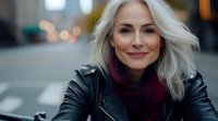 Confident mature woman with silver hair wearing black leather jacket and burgundy scarf smiling at camera outdoors with blurred urban background.
