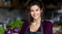 Young woman with dark hair wearing purple sweater and black apron smiling in kitchen with fresh vegetables including cabbage and greens, cooking preparation scene.
