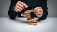 Business professional placing wooden block on unstable tower demonstrating risk management and strategic decision making in corporate environment.