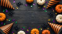 Halloween party decorations frame with orange pumpkins, striped party hats, black spiders and colorful confetti on dark wooden background with copy space.
