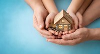 Family hands protecting wooden house model with coins on blue background symbolizing home insurance, mortgage protection and real estate investment security.