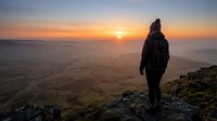 Hiker in dark jacket stands on rocky mountain peak watching golden sunrise over misty valley landscape below.
