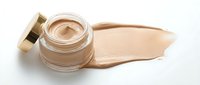 Open glass jar of beige foundation cream with golden cap and makeup smear on white background. Cosmetic product for skincare and beauty routine.
