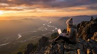Woman working on laptop while sitting on mountain peak at sunset, overlooking valley with winding river and distant hills, remote work and digital nomad lifestyle concept.