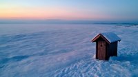 Rustic wooden ice fishing hut stands solitary on frozen lake surface against pastel winter sunset sky, creating serene Nordic atmosphere for outdoor adventure concepts.