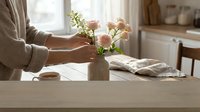Woman arranging fresh spring flowers in ceramic vase on wooden kitchen table. Cozy home interior with natural light creating warm atmosphere for lifestyle content.