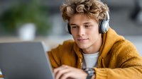 Young Caucasian man with curly blonde hair wearing headphones and yellow sweater working on laptop computer at home office desk for remote work.