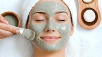 Young Asian woman with closed eyes and slight smile receiving green clay facial mask application with brush at spa. Skincare and beauty treatment procedure.