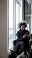 Happy African American businesswoman with gray curly hair using smartphone while sitting in modern office waiting area near large windows.