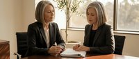 Two professional businesswomen in dark suits having meeting at conference table in modern office with natural lighting and documents.