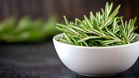 Fresh rosemary sprigs in white ceramic bowl on dark background with selective focus. Aromatic cooking herbs and natural food ingredients for Mediterranean cuisine.